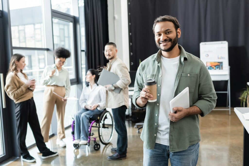 cheerful indian office worker holding coffee to go and tablet near diverse startup team in coworking